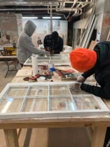 Two young men work on striping paint from old windows.