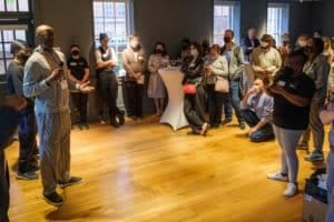 A room full of people stand in a museum space and listen to a speaker with a microphone.