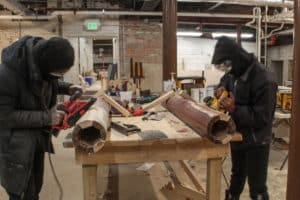 Two people wearing black sweatshirts use power tools to strip and sand columns.