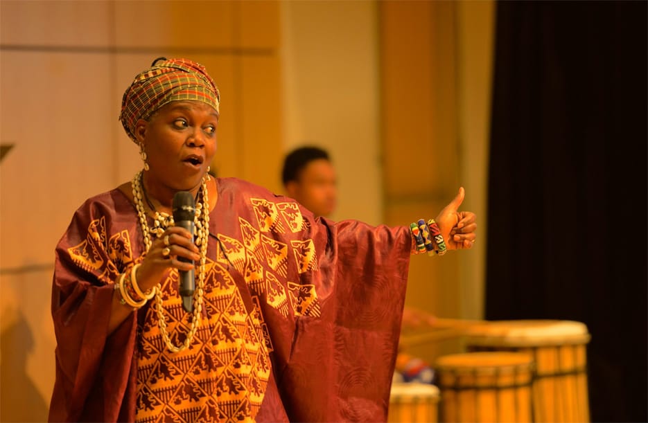 A woman in traditional African robes and a head scarf speaks with a microphone on a stage.