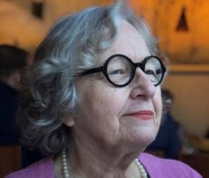 Romaine Sommerville has black glasses with round rims, a pearl necklace, and a purple shirt.