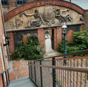 The Peale's garden has a brick pathway, green bushes, and a wooden and metal staircase leading down from a second floor room.
