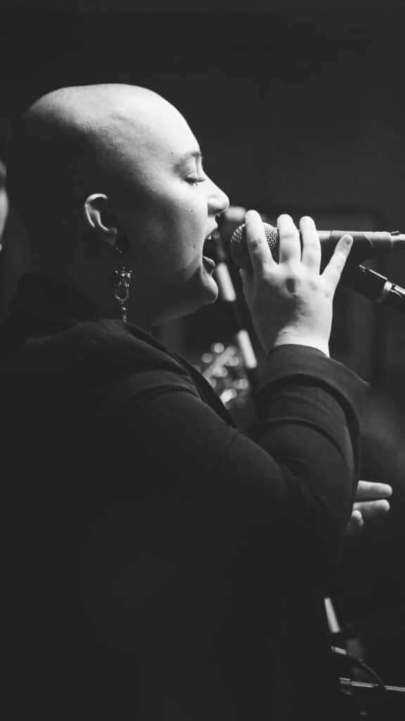 A woman with a black sweater and long earrings sings into a microphone.
