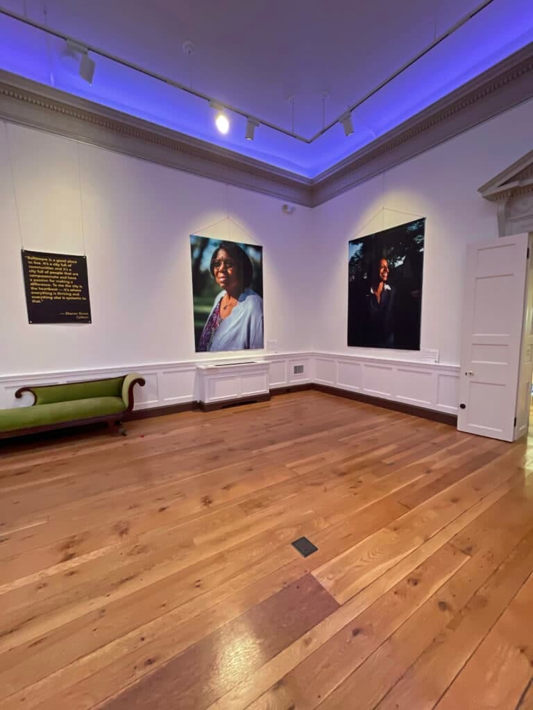 An exhibition gallery with hard wood floors and large portraits on the white walls. A blueish light illuminates the ceiling.