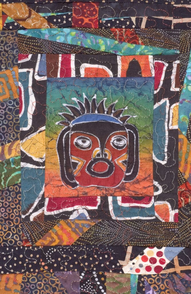 A detail of a quilt that shows a face in the style of an Aztec mask in brown, red, and blue in the center.