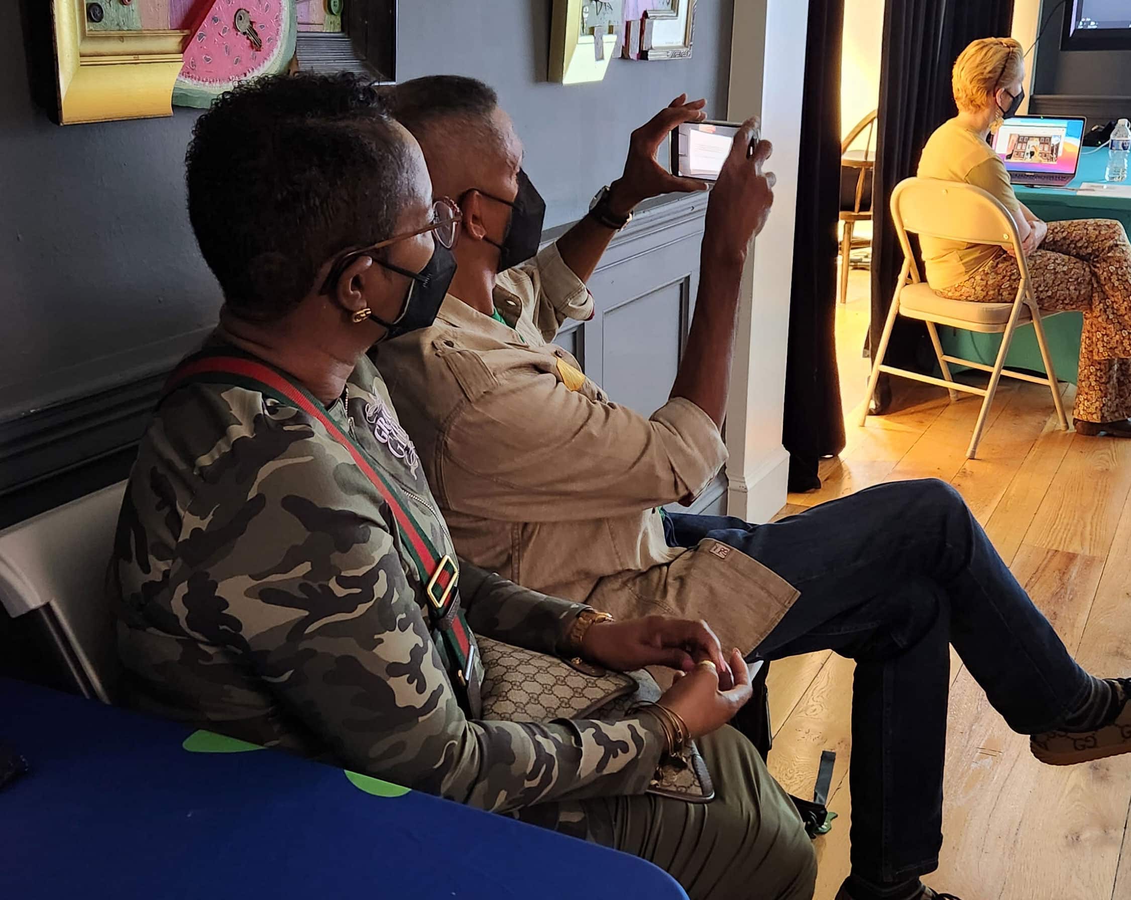 Two people sit and listen to a speaker in a gallery space with dark, gray walls. One person holds a smartphone to take a picture.