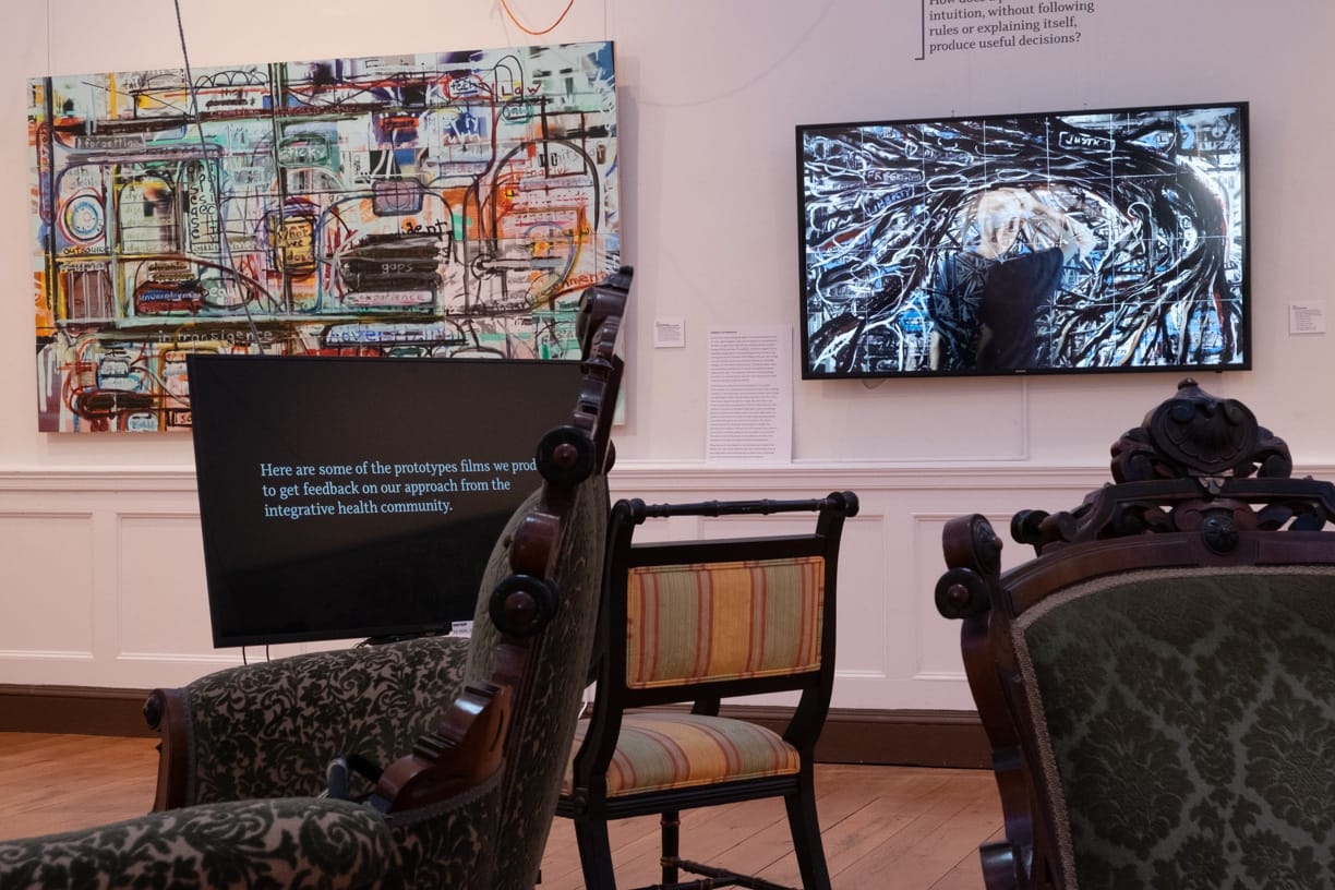 A gallery space with modern art on the walls and vintage looking chairs in the middle of the room.