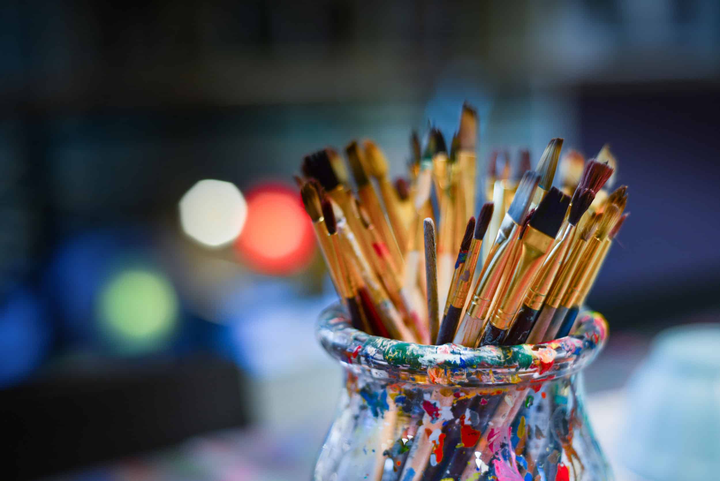 A glass vase covered with paint, full of used paint brushes.