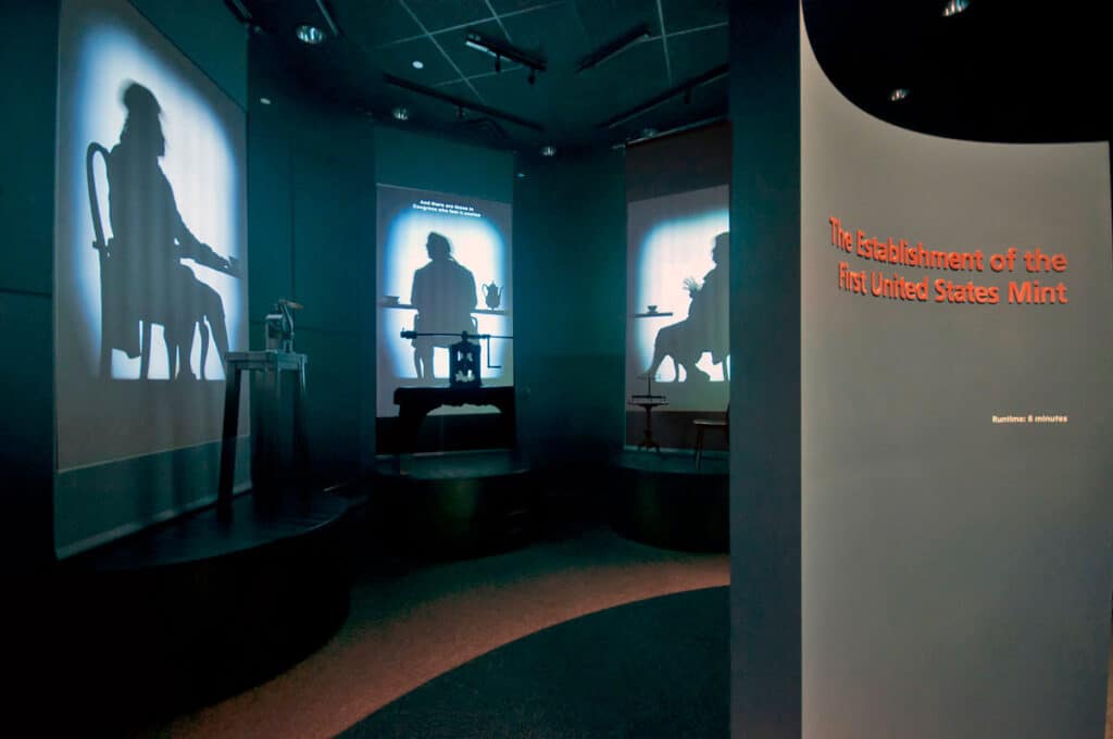 A curved wall that peeks into another room that shows shadowy figures of people projected on a wall.