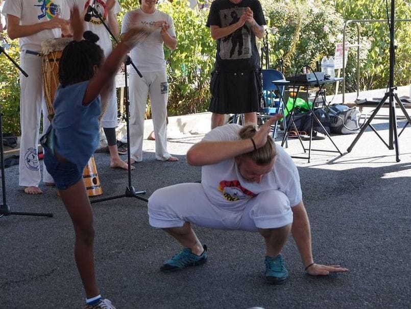 An man wearing white squats down while dancing with a group of people outside.