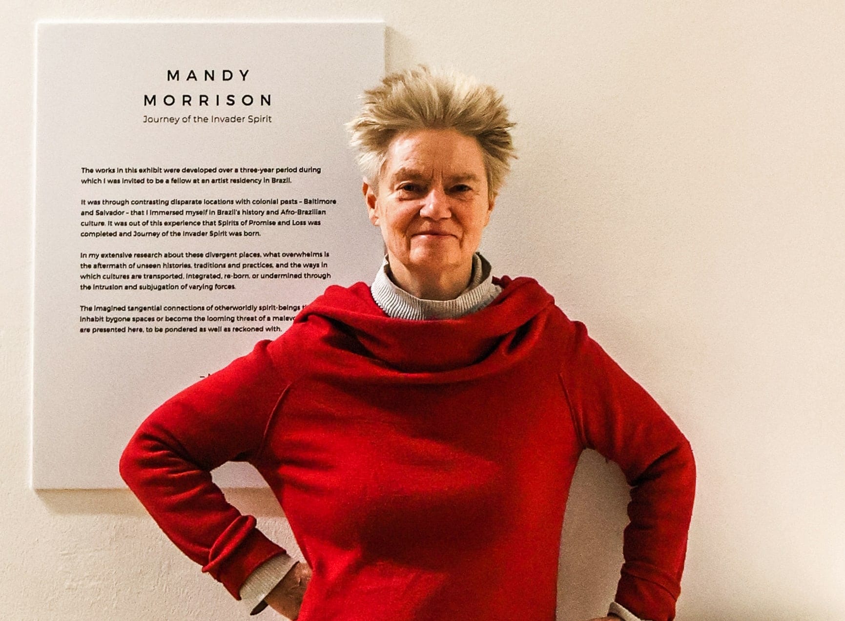 Mandy Morrison wears a bright red sweater and stands in front of a panel with text on it.