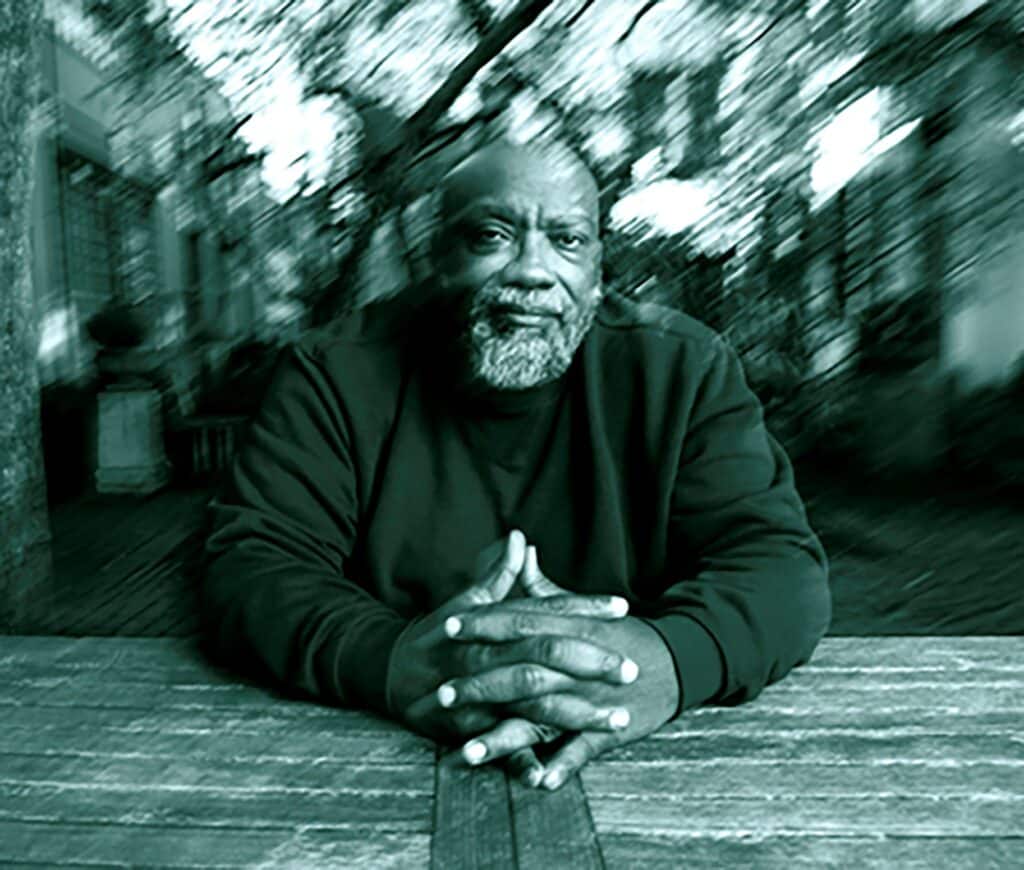 Fred Moten has a black shirt and folds his hands on a table. A blurry background is behind him.