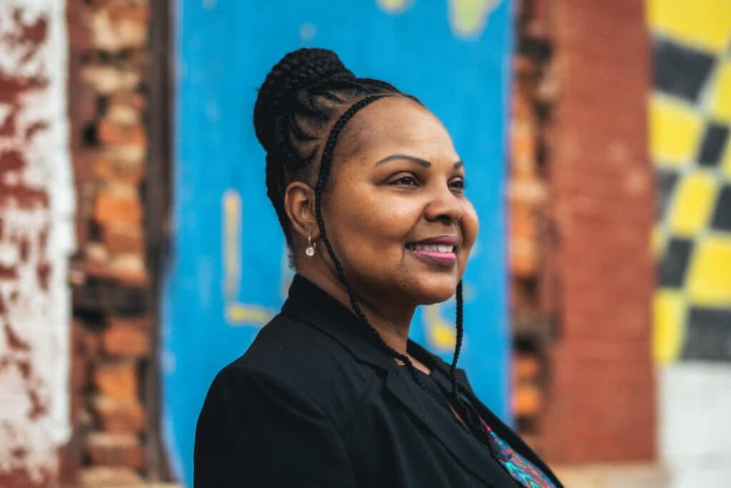 A woman with a black blazer and her hair in a bun on top of her head stands in profile and is photographed in front of a blue wall.