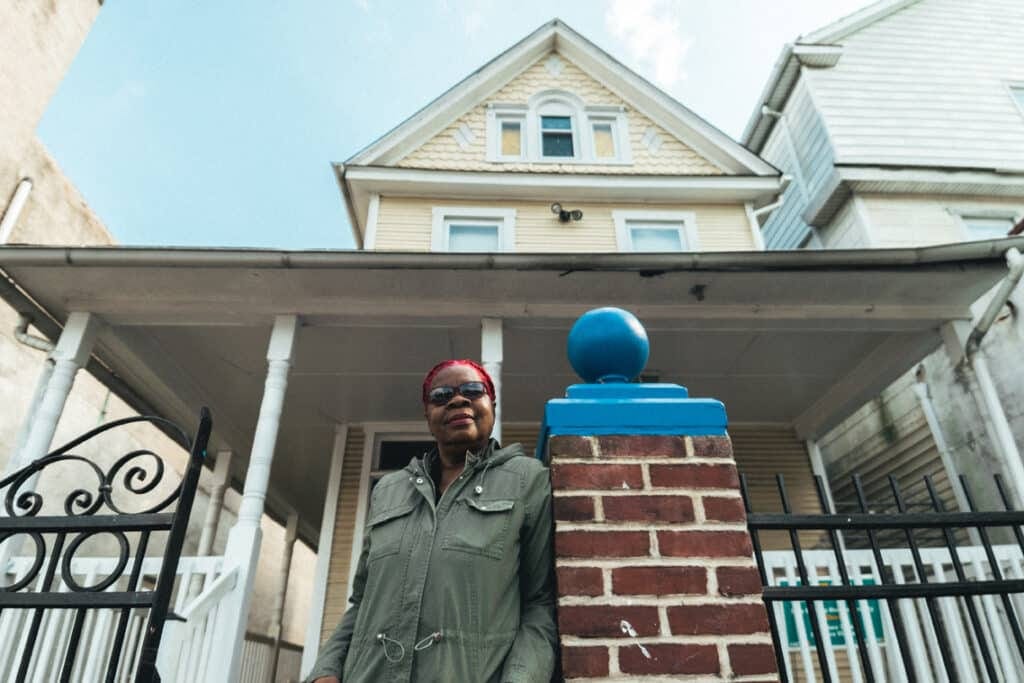 A woman with bright pink hair leans against a brick pillar leading up to the front porch of a house.
