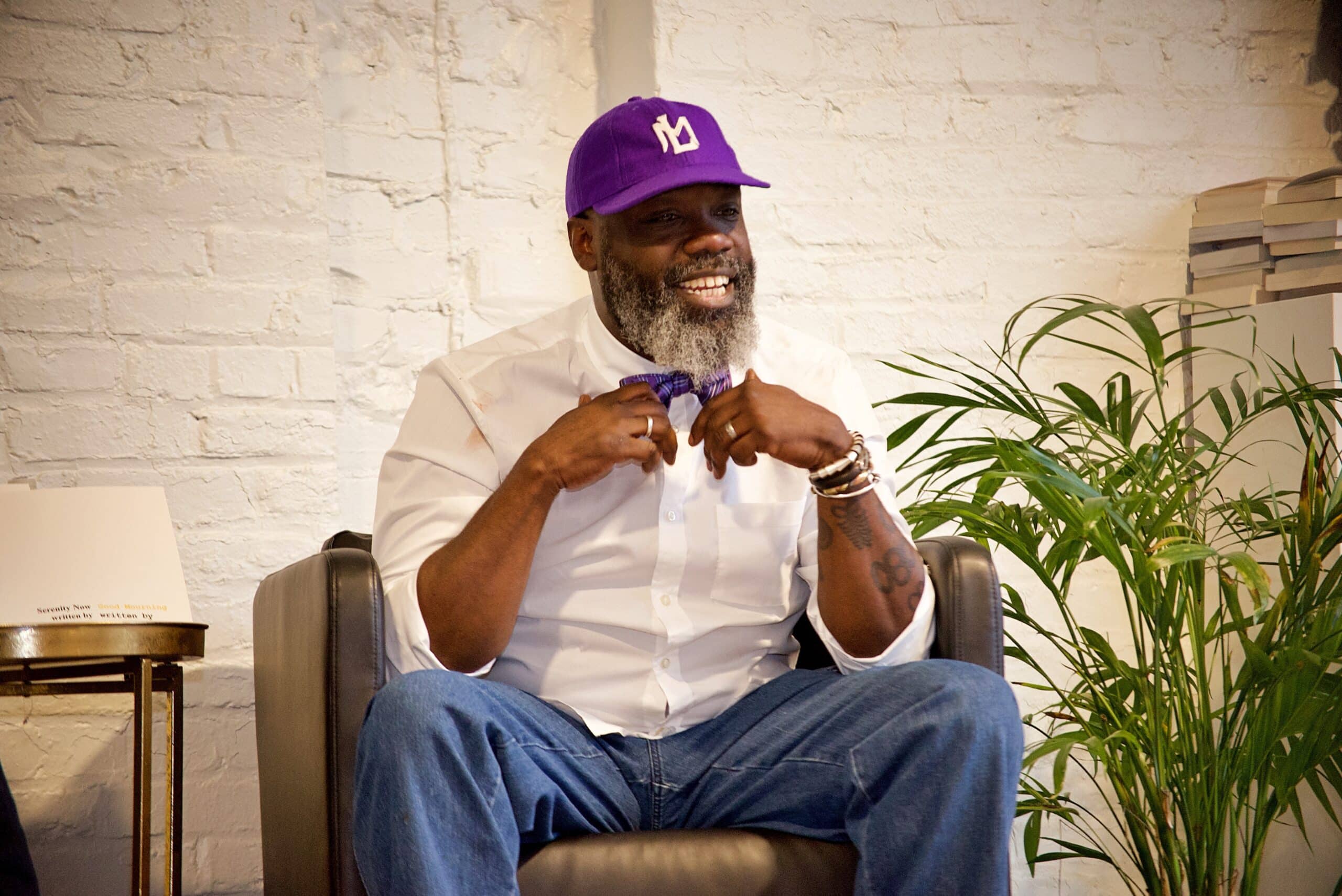 A man with a purple hat and white shirt sits in a chair and talks with a crowd.