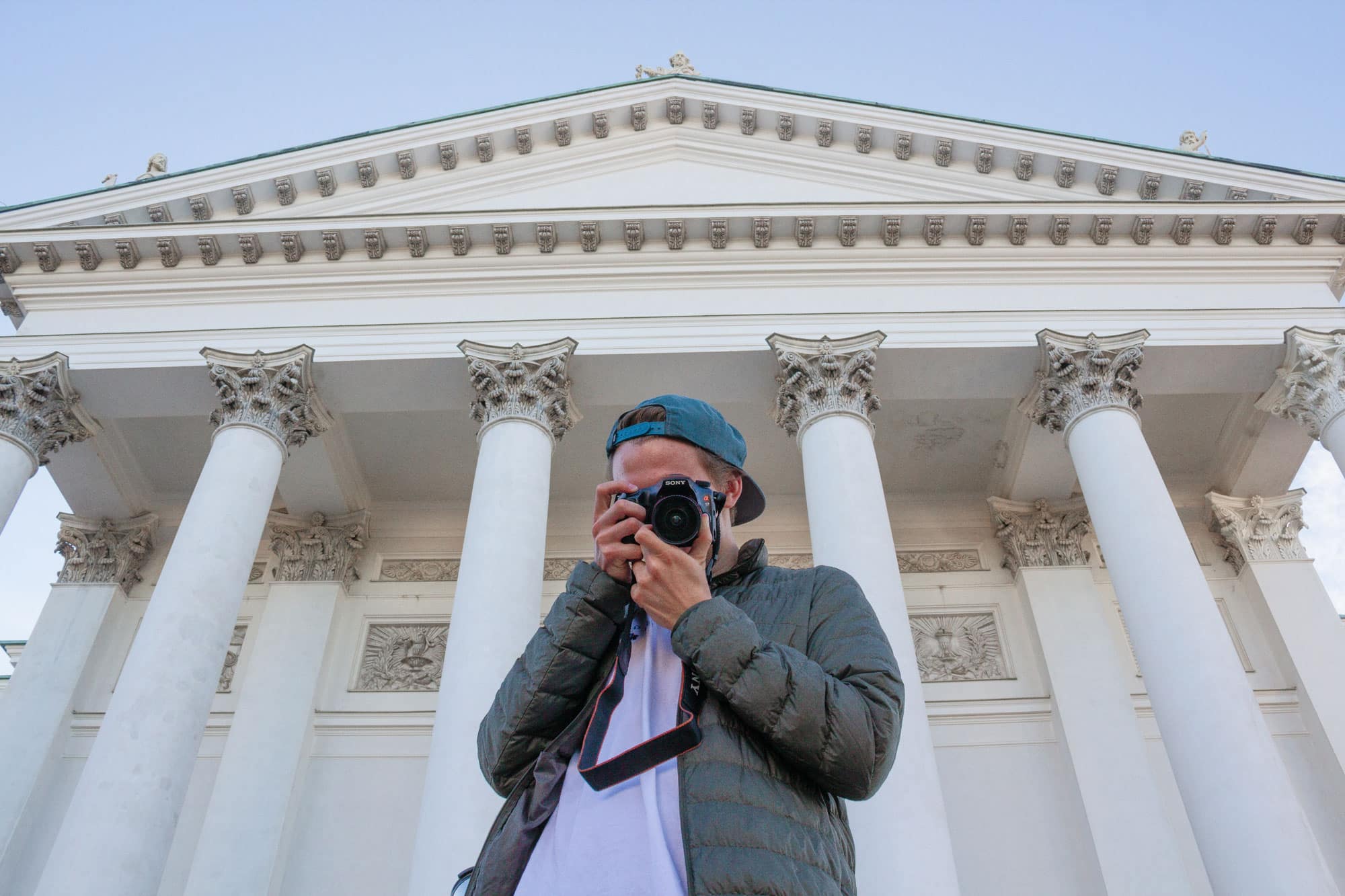 A man holds a camera up to his eye and snaps a photo while standing in front of a classical building with columns.