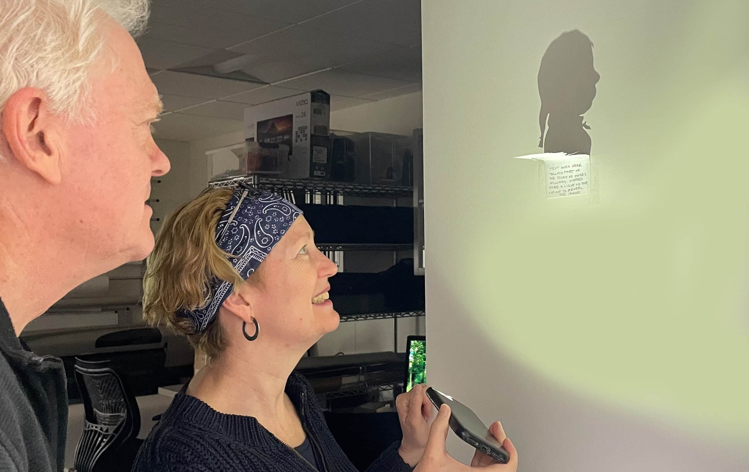 A woman shines a light on a piece of paper that casts a shadow on a wall.