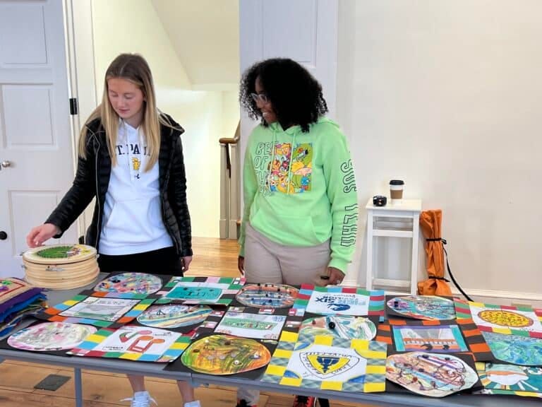 Two students look over colorful artwork that is laid out on a table in a museum space.