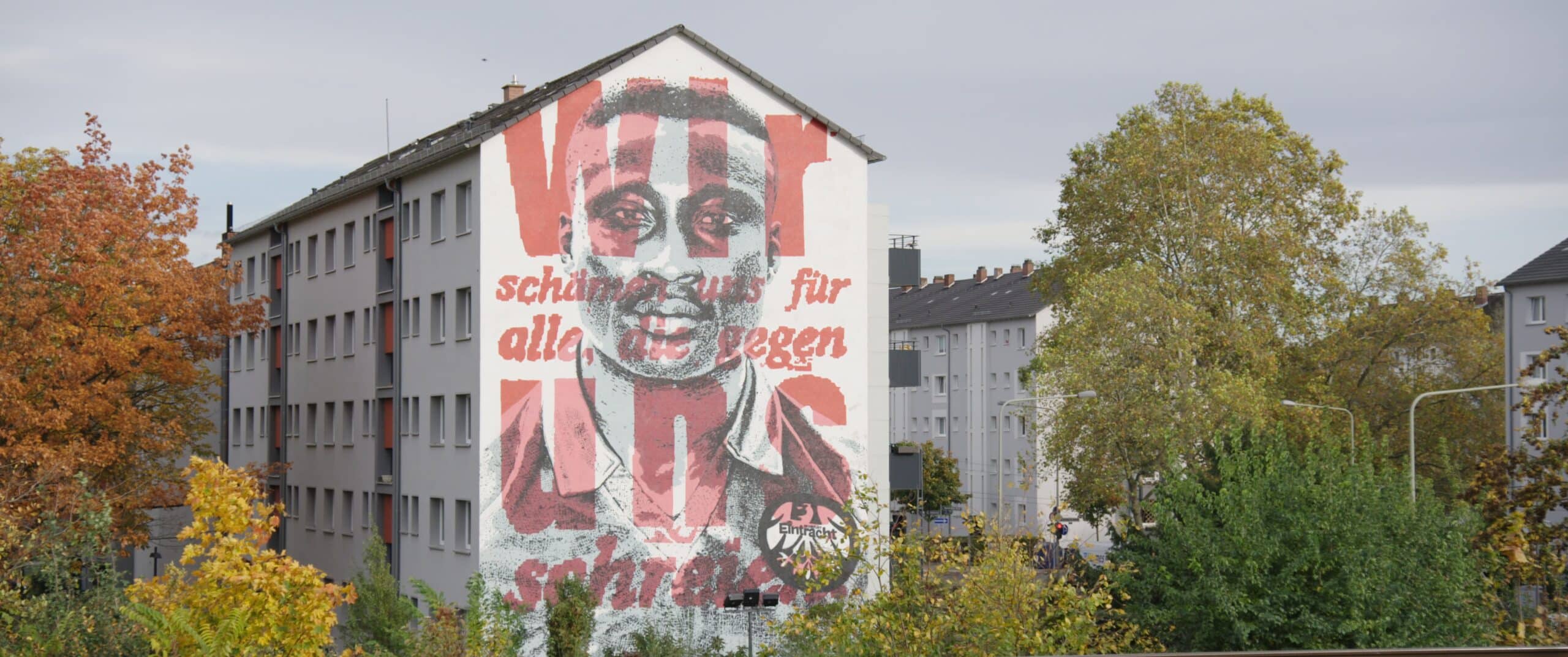 A warehouse or factory with a large mural of a person's face on it and words in German laid over top.