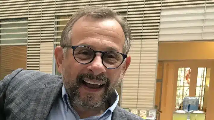 A man with round-rimmed glasses smiles broadly in a museum setting.