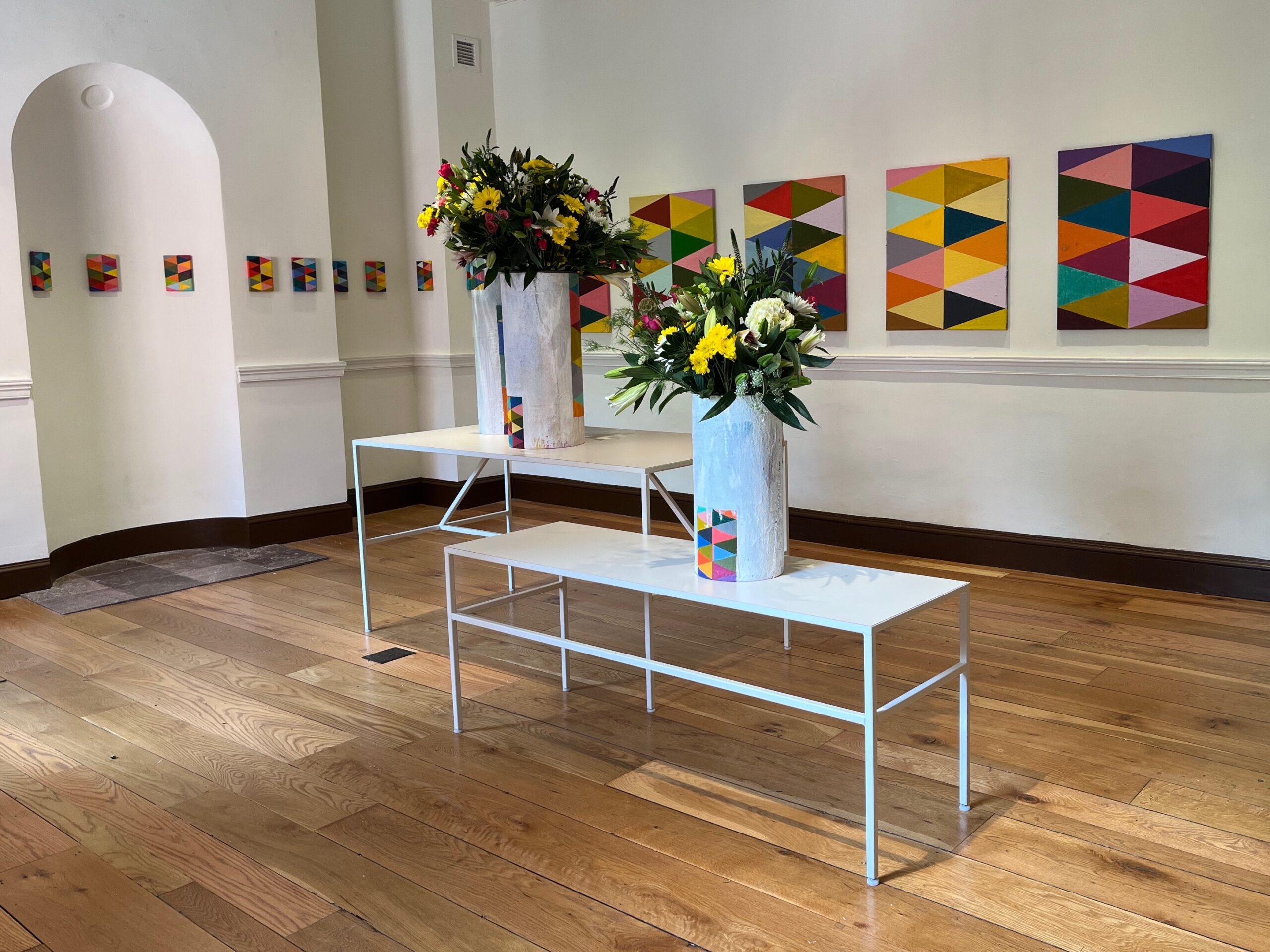 A museum gallery with contemporary looking white tables and two vases of flowers on them.