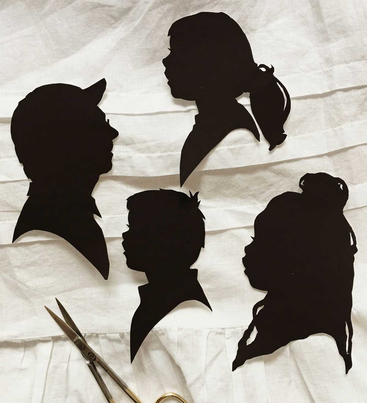 Four silhouettes on black paper that rest on a white piece of cloth.