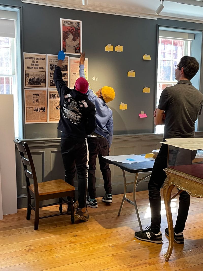 Three people install a museum exhibition in a room with gray walls.