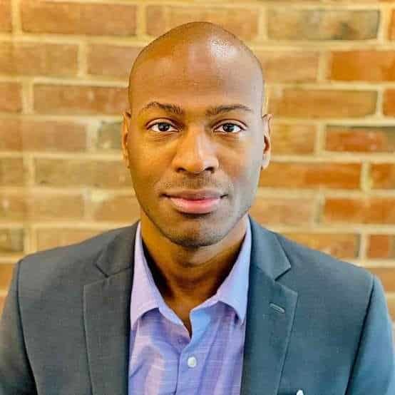 An man with a purple dress shirt and gray jacket stands in front of a brick wall.