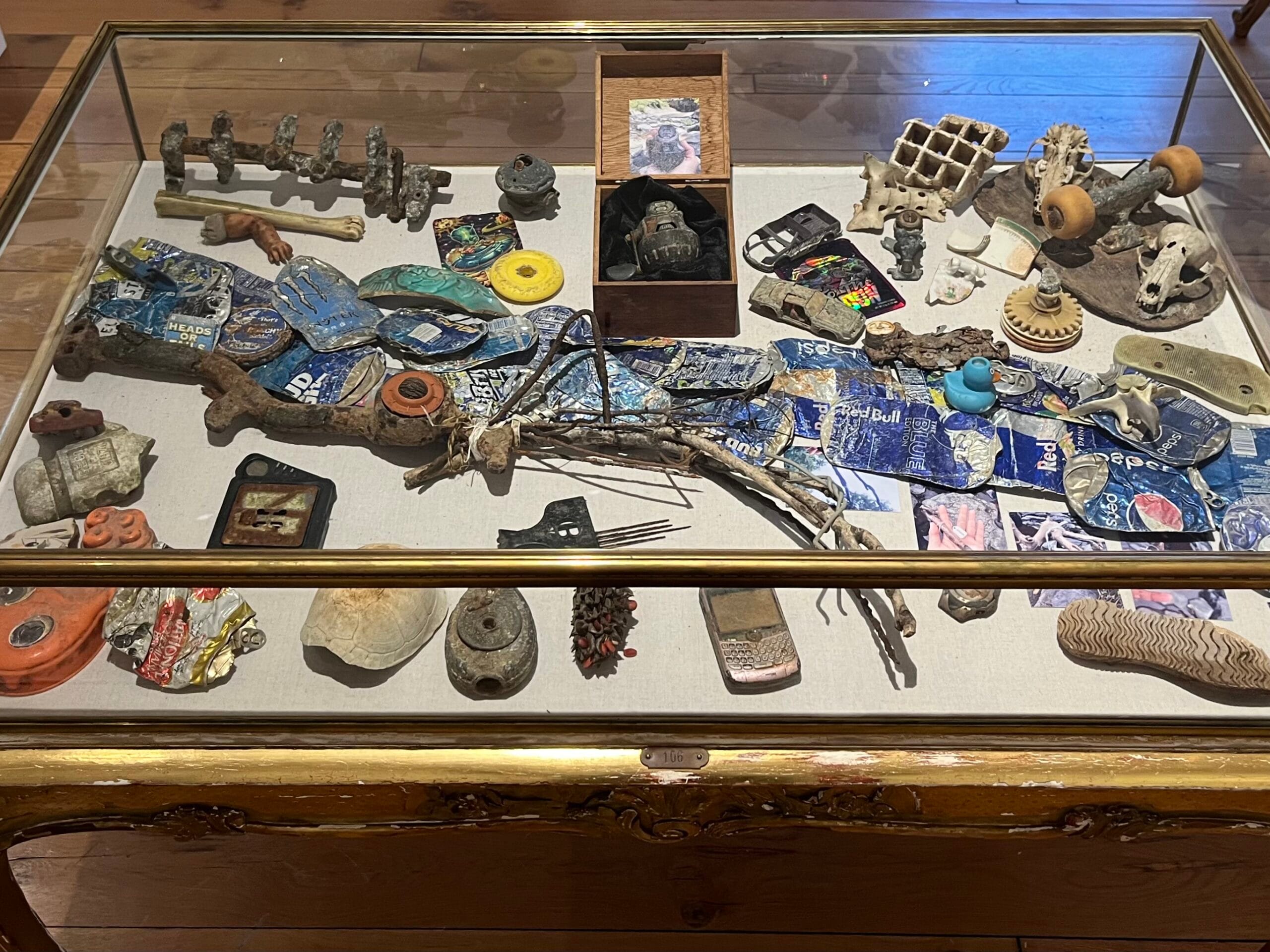 An exhibition case filled with trash--objects found on a river bank, like a skateboard, a shoe, and an old phone.