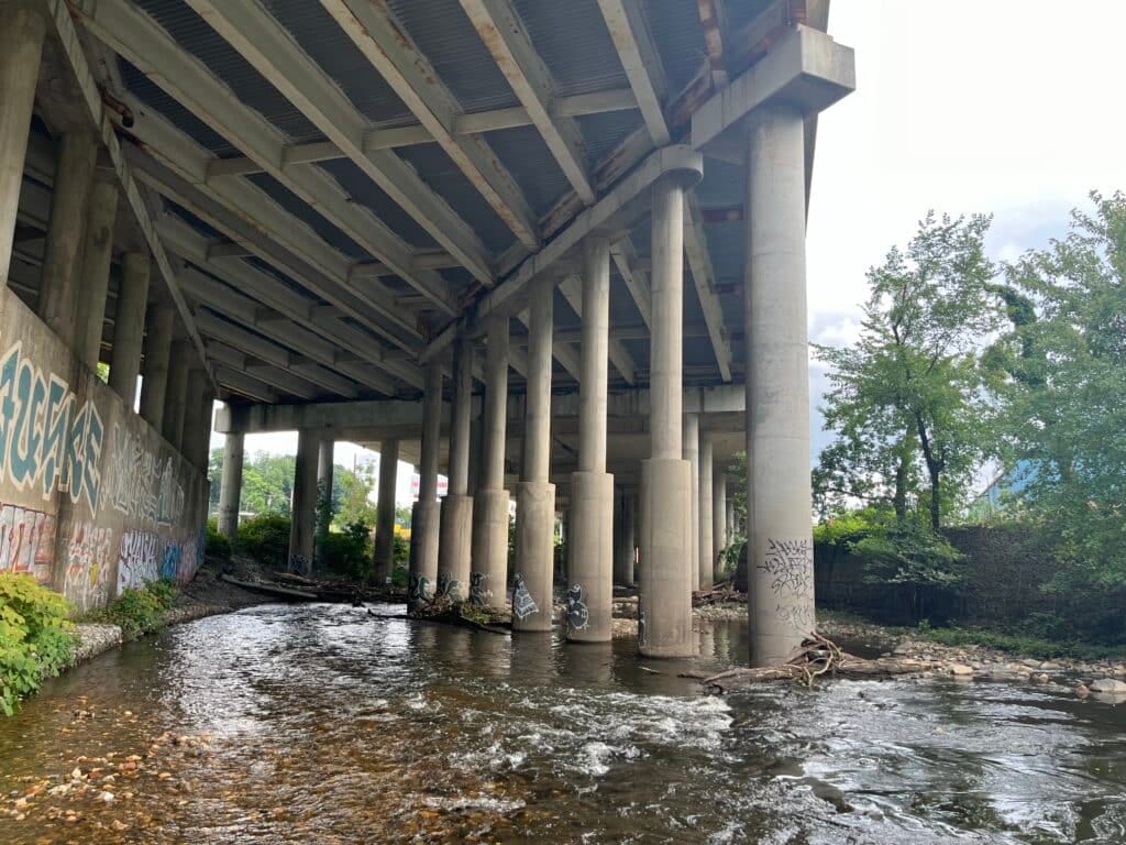 A stream babbles underneath an urban bridge with concrete supports.