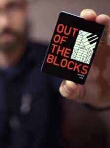 A man holds up a deck of cards that says, Out of the Blocks.