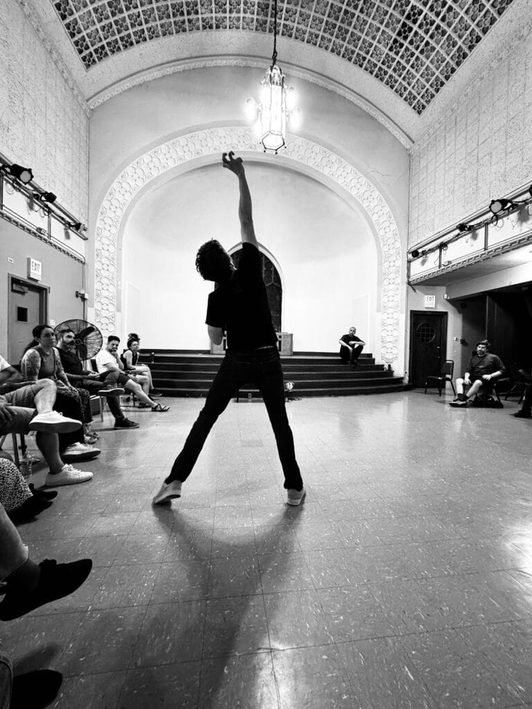 A man in black dances with his feet spread apart and one arm in the air.