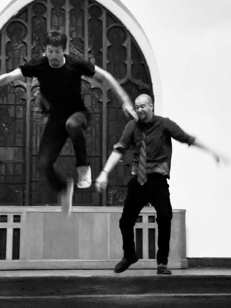 Two men jump from a stage. One man is in mid-air and raises his legs.