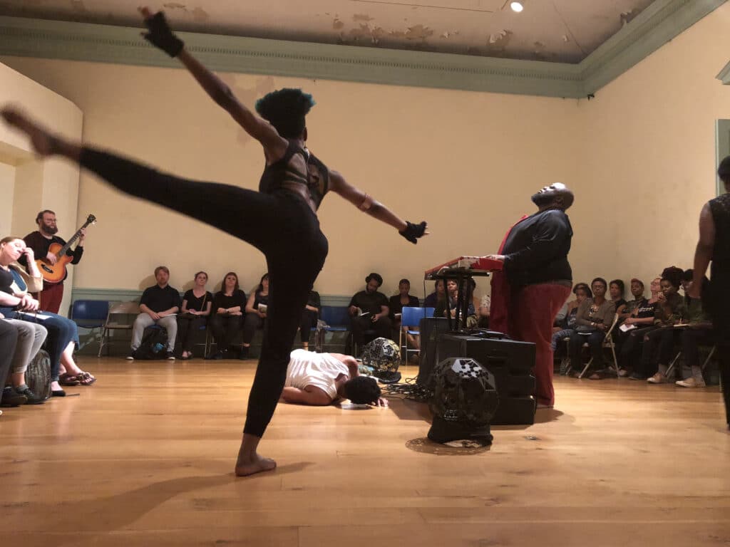 Dancer mid high kick on a wooden stage, audience seated along the back wall, guitarist visible on the left and a keyboard performer on the right.