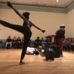 Dancer mid high kick on a wooden stage, audience seated along the back wall, guitarist visible on the left and a keyboard performer on the right.