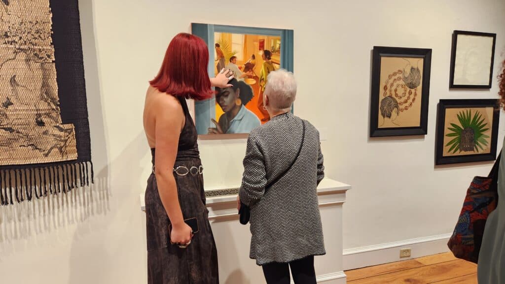 Two visitors in an art gallery study a large framed painting together, with more artworks on the wall to the right and a woven piece on the left side of the image.