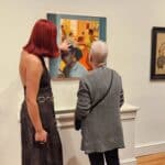 Two visitors in an art gallery study a large framed painting together, with more artworks on the wall to the right and a woven piece on the left side of the image.