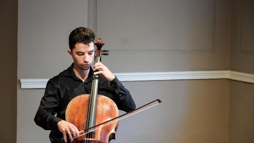 Man in a black shirt playing a cello in a plain room, bow gliding across strings.