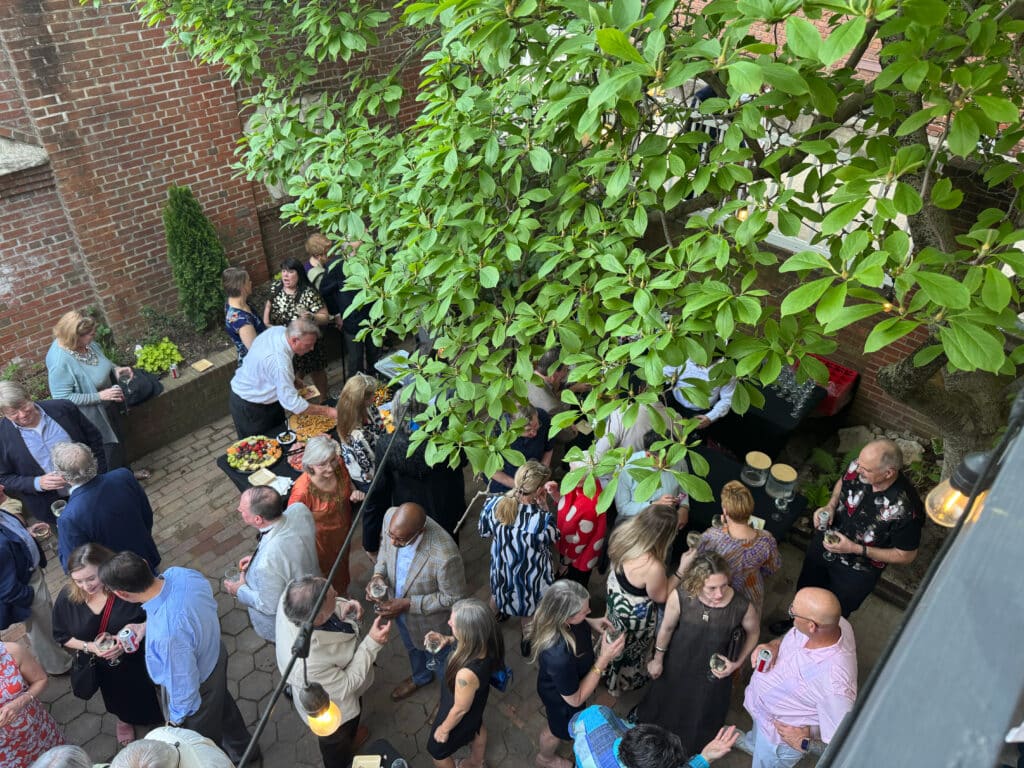 Crowd of guests at an outdoor courtyard party by a brick wall, with tables of food and drinks.