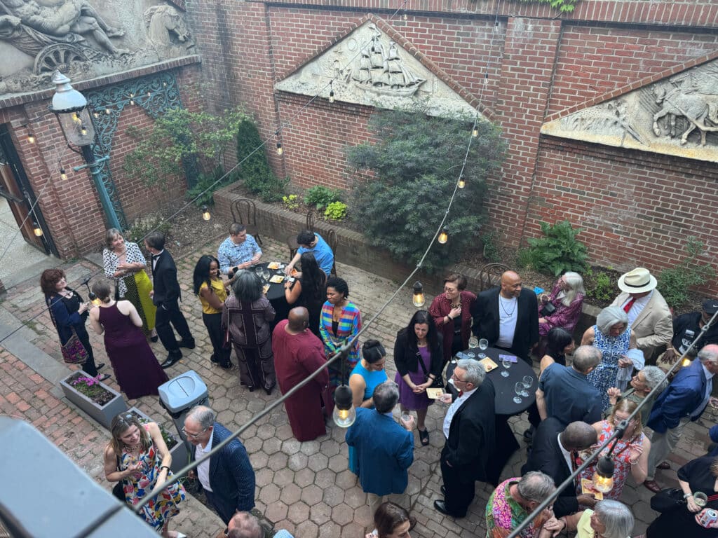 People in colorful outfits mingle around tables with wine glasses in a brick-walled courtyard under string lights at an outdoor party.