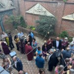 People in colorful outfits mingle around tables with wine glasses in a brick-walled courtyard under string lights at an outdoor party.