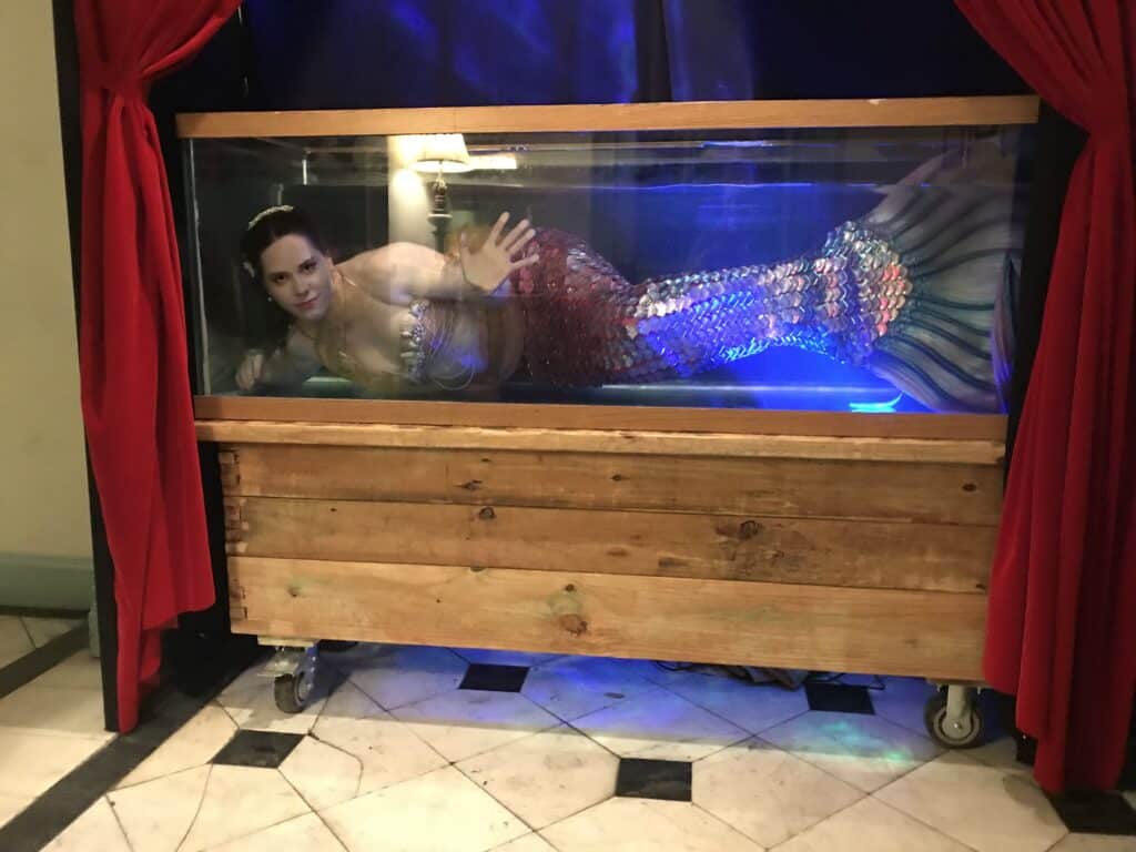 A mermaid prop inside a glass tank on a wheeled wooden frame, with red theater curtains on both sides.