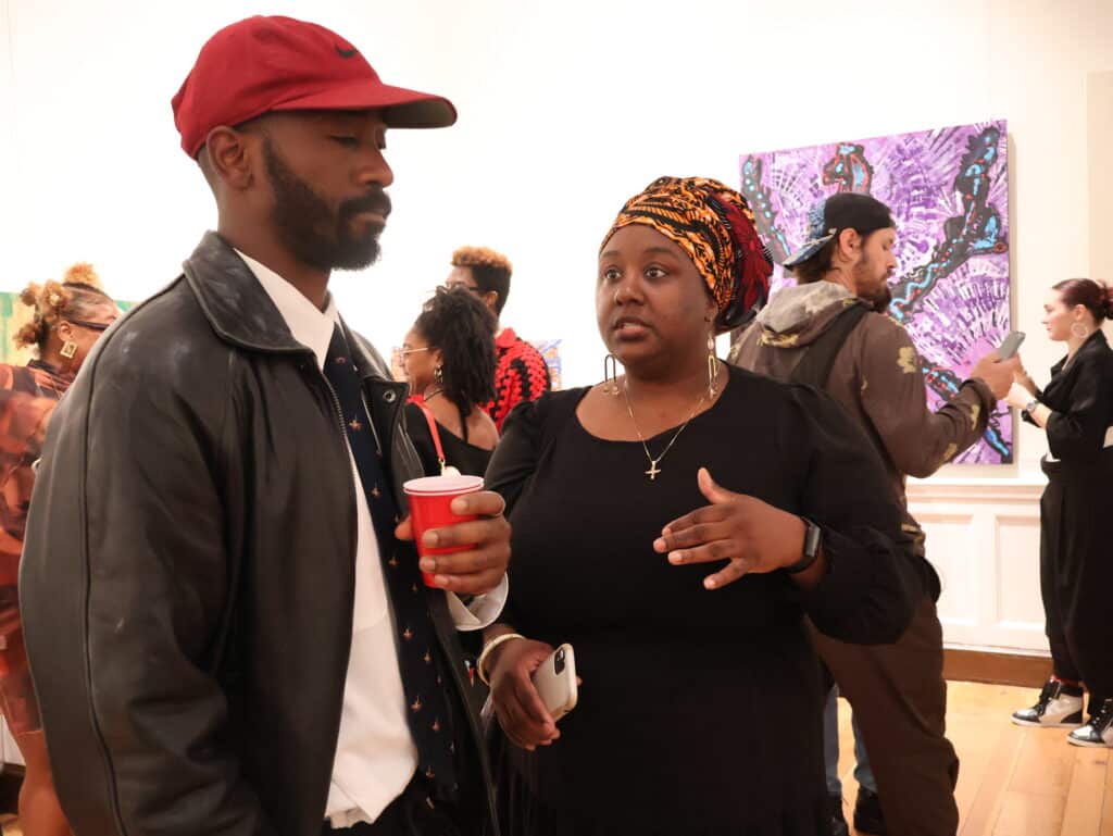 Two people conversing at an art gallery, with colorful paintings in the background; man in a red cap and leather jacket, woman wearing a patterned head wrap hold drinks nearby.