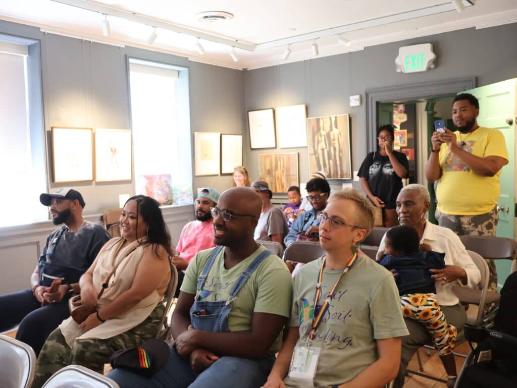 D diverse audience seated in a gallery, with framed art on gray walls, while a man in a yellow shirt photographs the event.