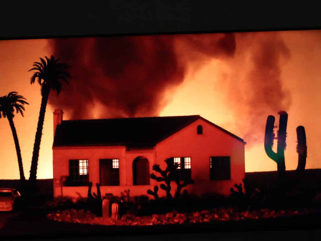 A house engulfed in orange flames with dark smoke, palm trees silhouettes in the foreground at night.