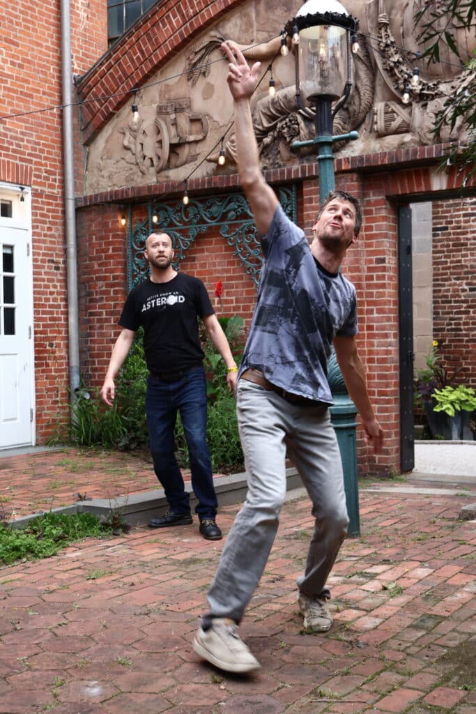 Two men outdoors beside a brick building with string lights; the man in the foreground reaches up toward a hanging bulb while the other watches.