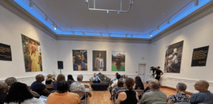 Audience seated in a gallery-style room facing a speaker at a small table with portraits on the walls behind them.