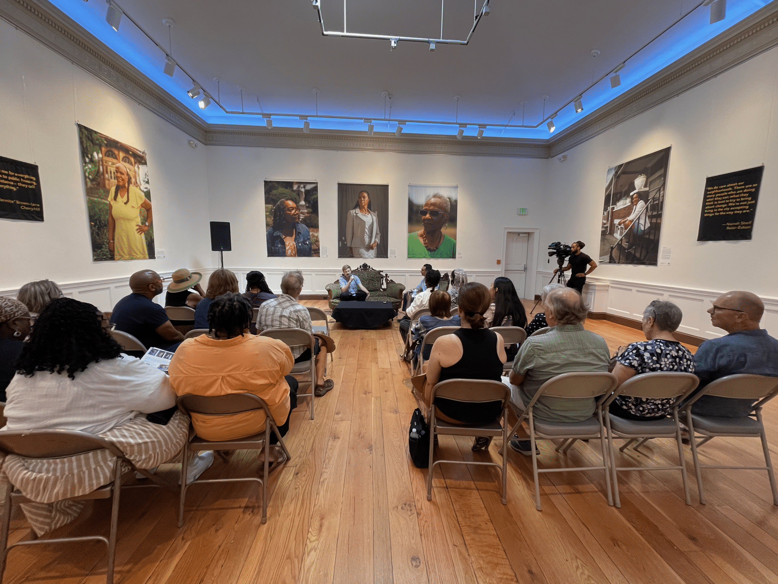 Audience seated in a gallery-style room facing a speaker at a small table with portraits on the walls behind them.