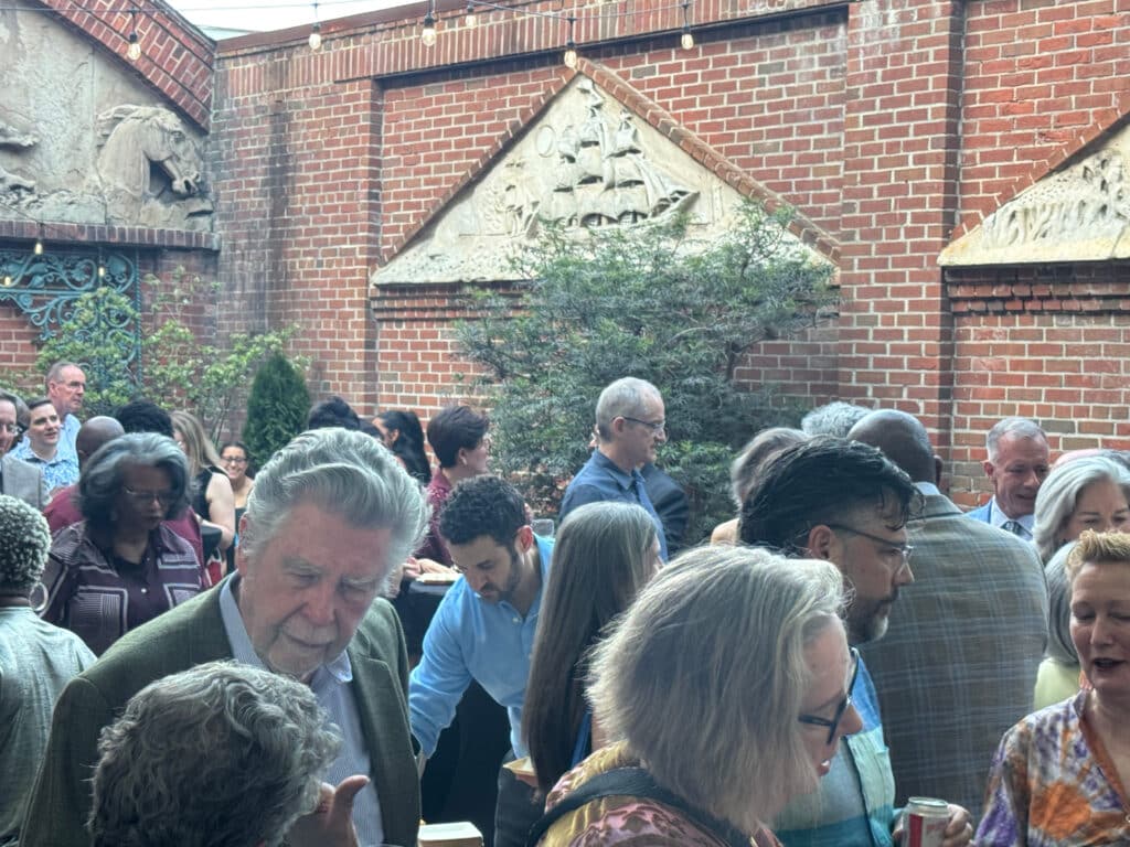 Crowd of people mingling at an outdoor event in a brick courtyard with string lights overhead.