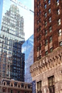 City street with tall glass skyscrapers reflecting neighboring buildings beside a brick and stone historic facade.
